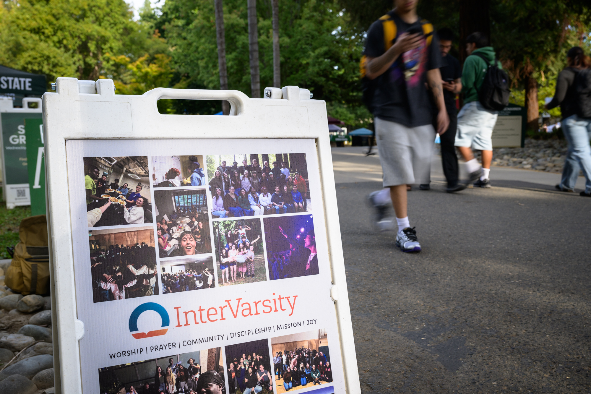 An InterVarsity A-frame sign sits next to a crowded walking path on campus; it showcased many photos of the chapter having fun in a variety of locations.