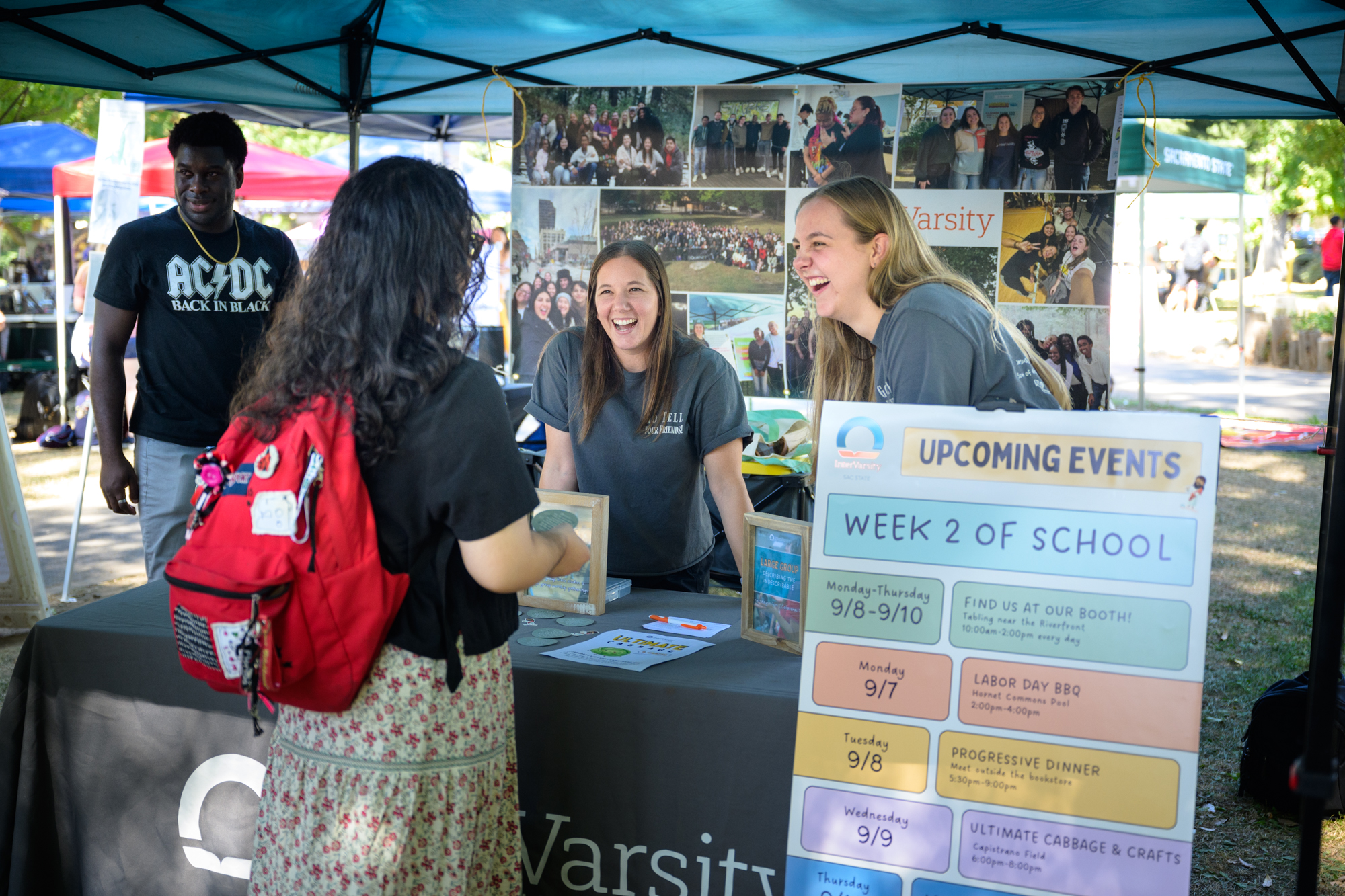 A staff worker and student stand behind an InterVarsity table, smiling and laughing at something a student, who is in front of the table, said. The list of the week’s events are listed on a poster nearby.
