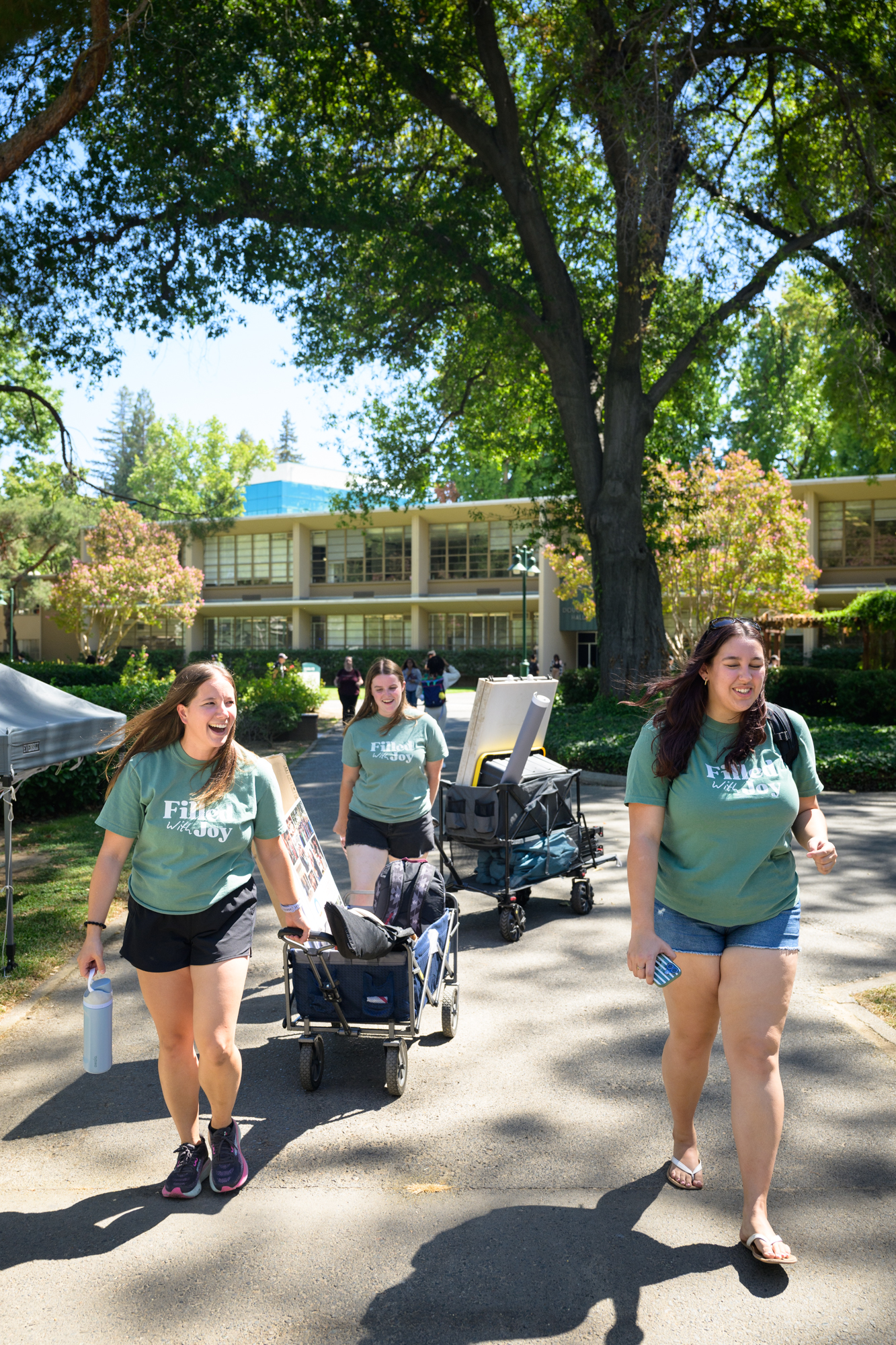 A student walks next to two staff workers as they each pull a wagon overflowing with supplies for tabling.