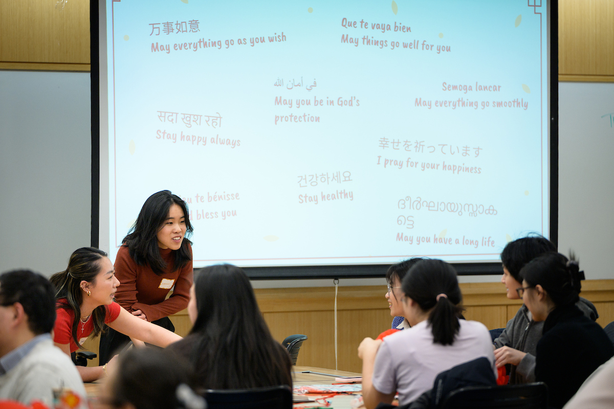 Hannah and other students gather at a table to make blessing lanterns with a screen behind them projecting blessing messages in nine languages