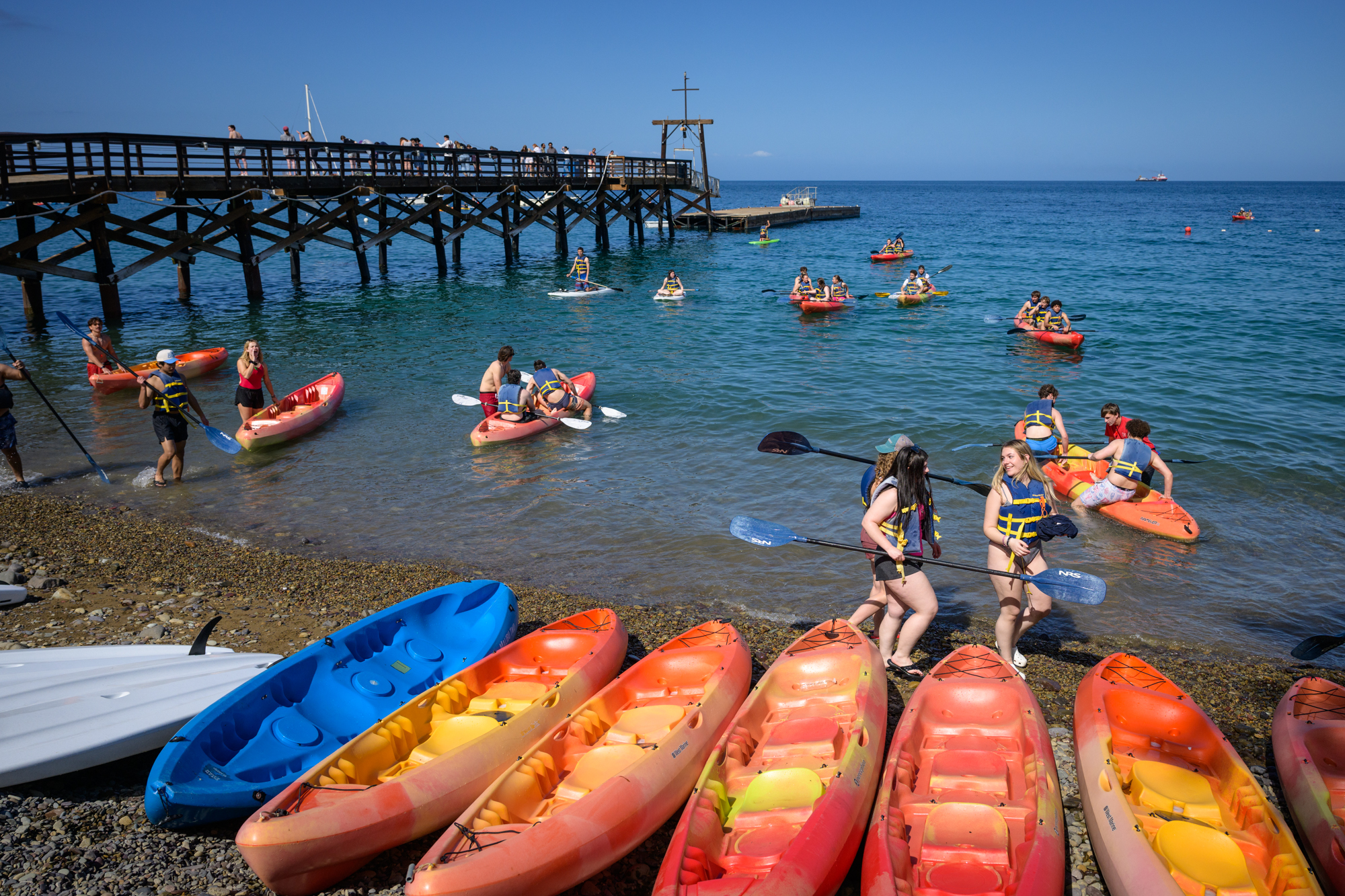 3 students walk on the rocky beach in life jackets. Several kayaks are in the foreground and many students are rowing kayaks in the ocean behind them.