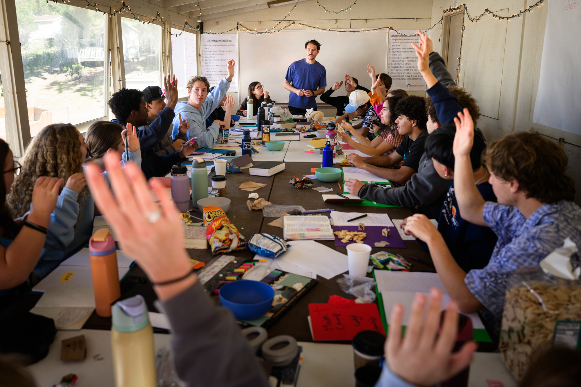 Track time - Students sit around a table littered with snacks and reference materials and water bottles. Students raise their hands to answer questions from the leader up front.
