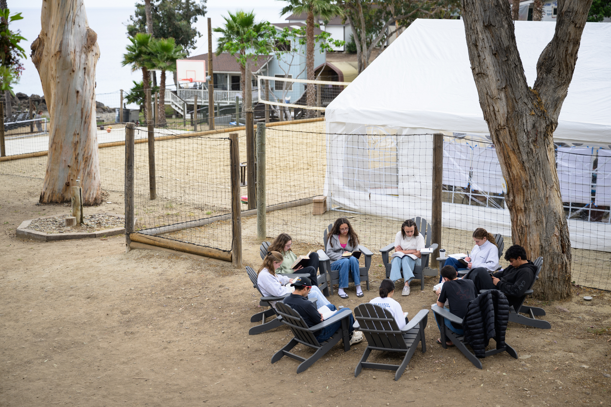 students sitting together in circle outside studying the Bible. The volleyball court, basketball court, and ocean are seen in the background.