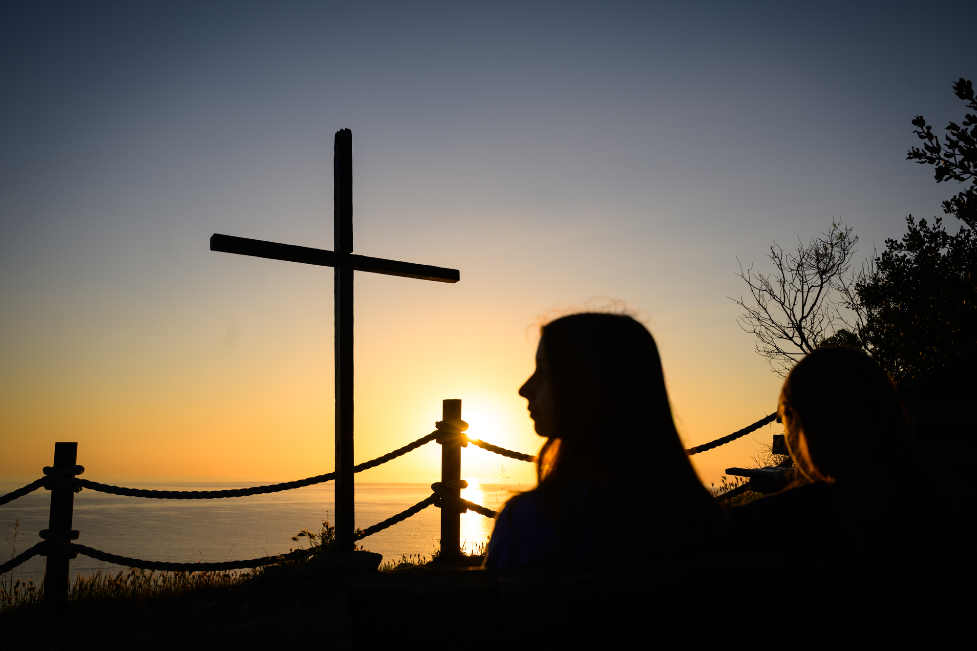 two students looking across the ocean standing on the hill with the cross silhouetted against the sunrise