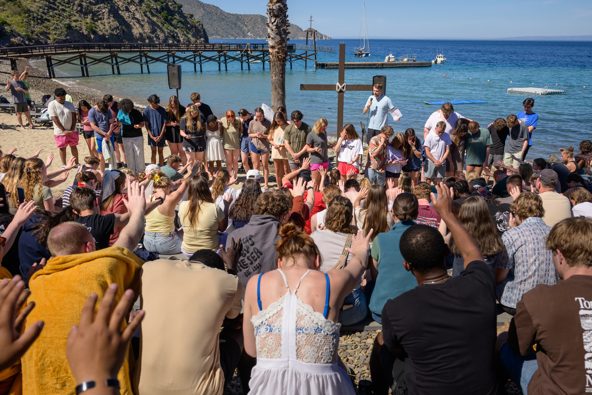 In the amphitheater in front of the ocean a crowd of students extends their hands in prayer towards the 29 students about to be baptized.