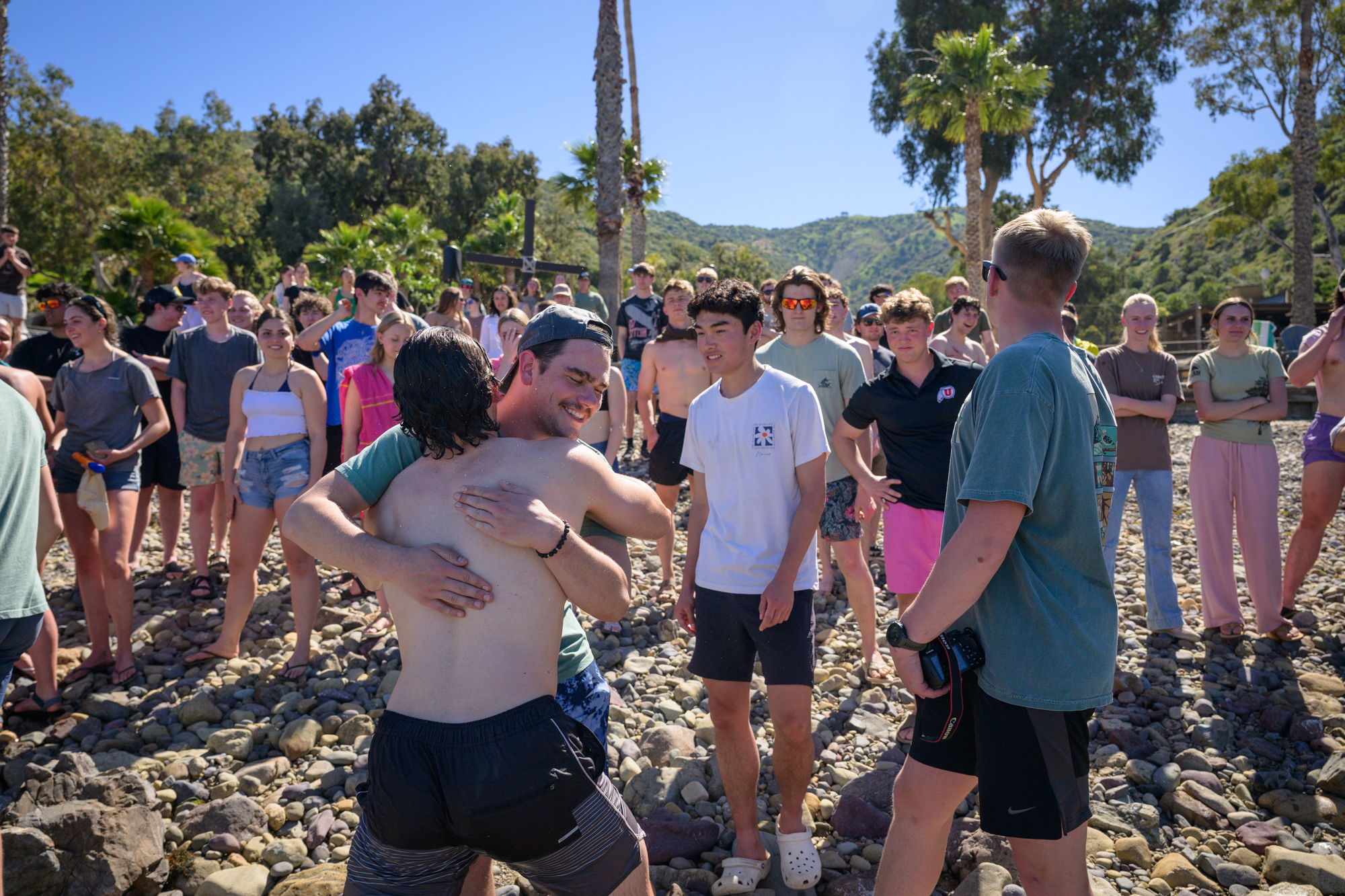 two men embrace on the beach after one comes out of the water after being baptized