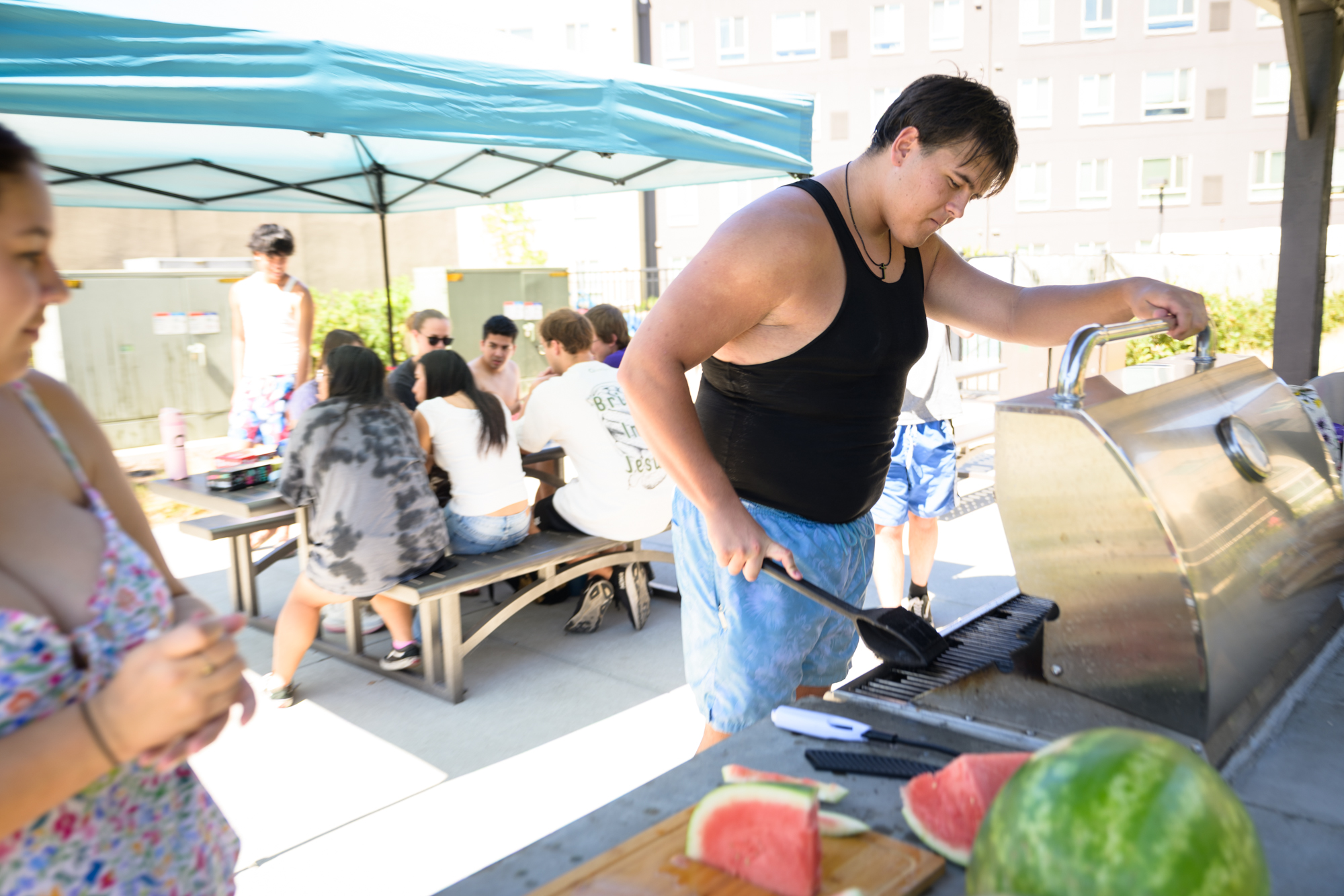 An InterVarsity student scrapes a BBQ, prepping it to grill, as a table fills with other students behind him.