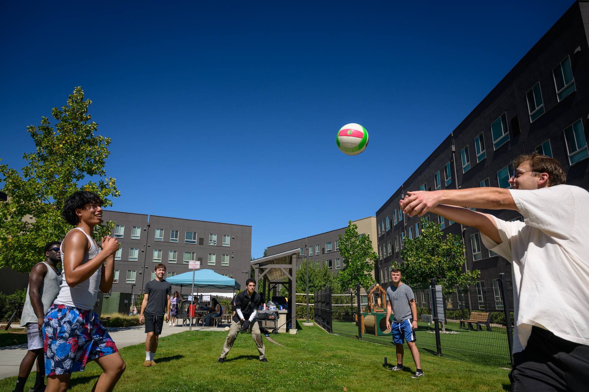 A student in the foreground hits a volleyball up into the air as several other students stand in a circle, ready to hit it back. The InterVarsity awning and BBQ pit are behind them, and a large dorm building sits in the background.