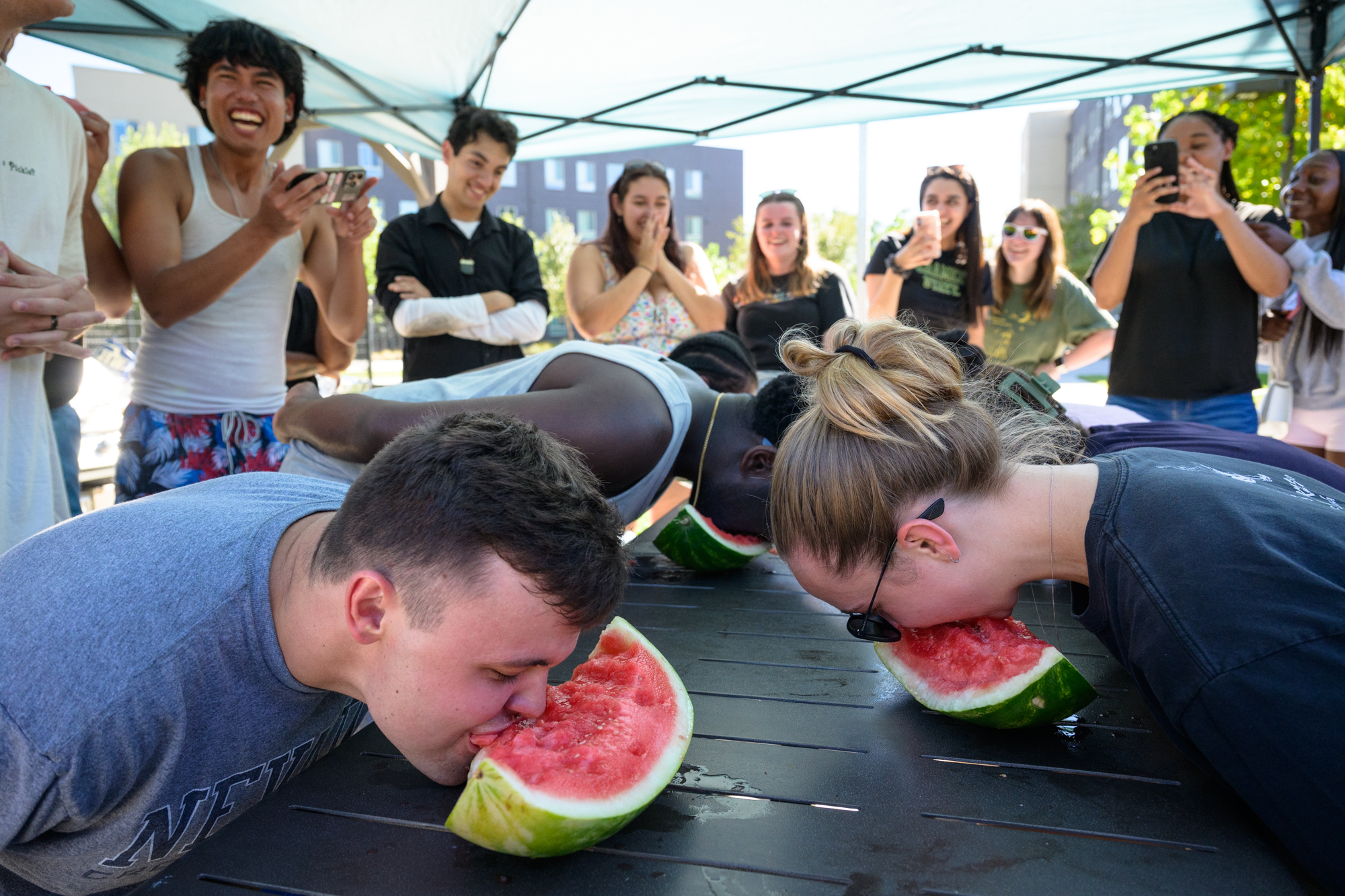Six students lean over large rinds of watermelon, eating with their hands behind their back in a contest of speed; many other students stand behind them laughing and cheering them on in disbelief.