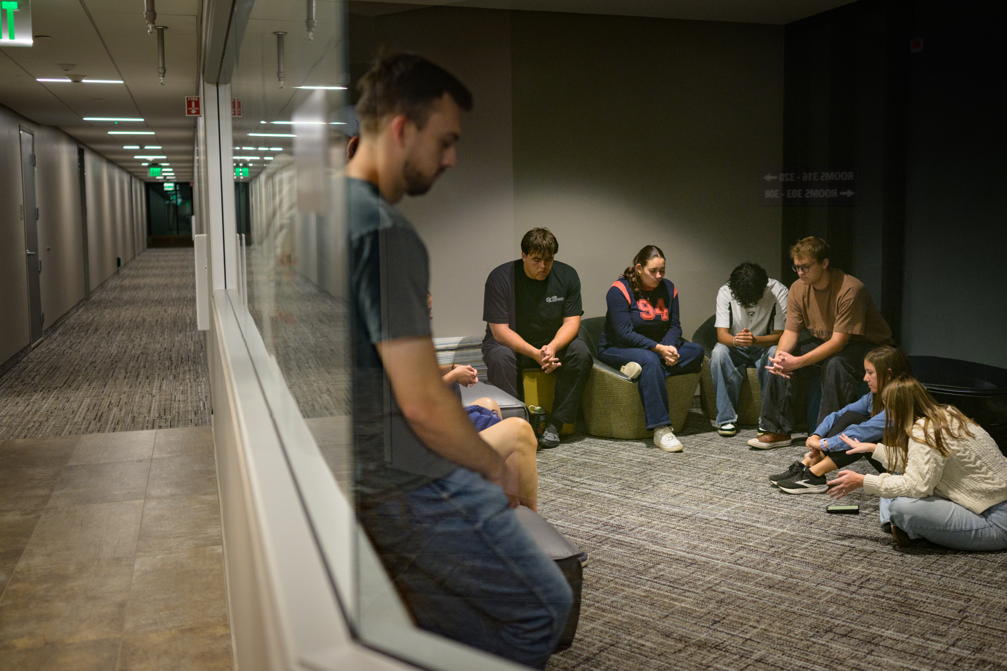 Students sit in a dorm lounge with their hands folded in prayer; outside is the hallway to all the dorm rooms.