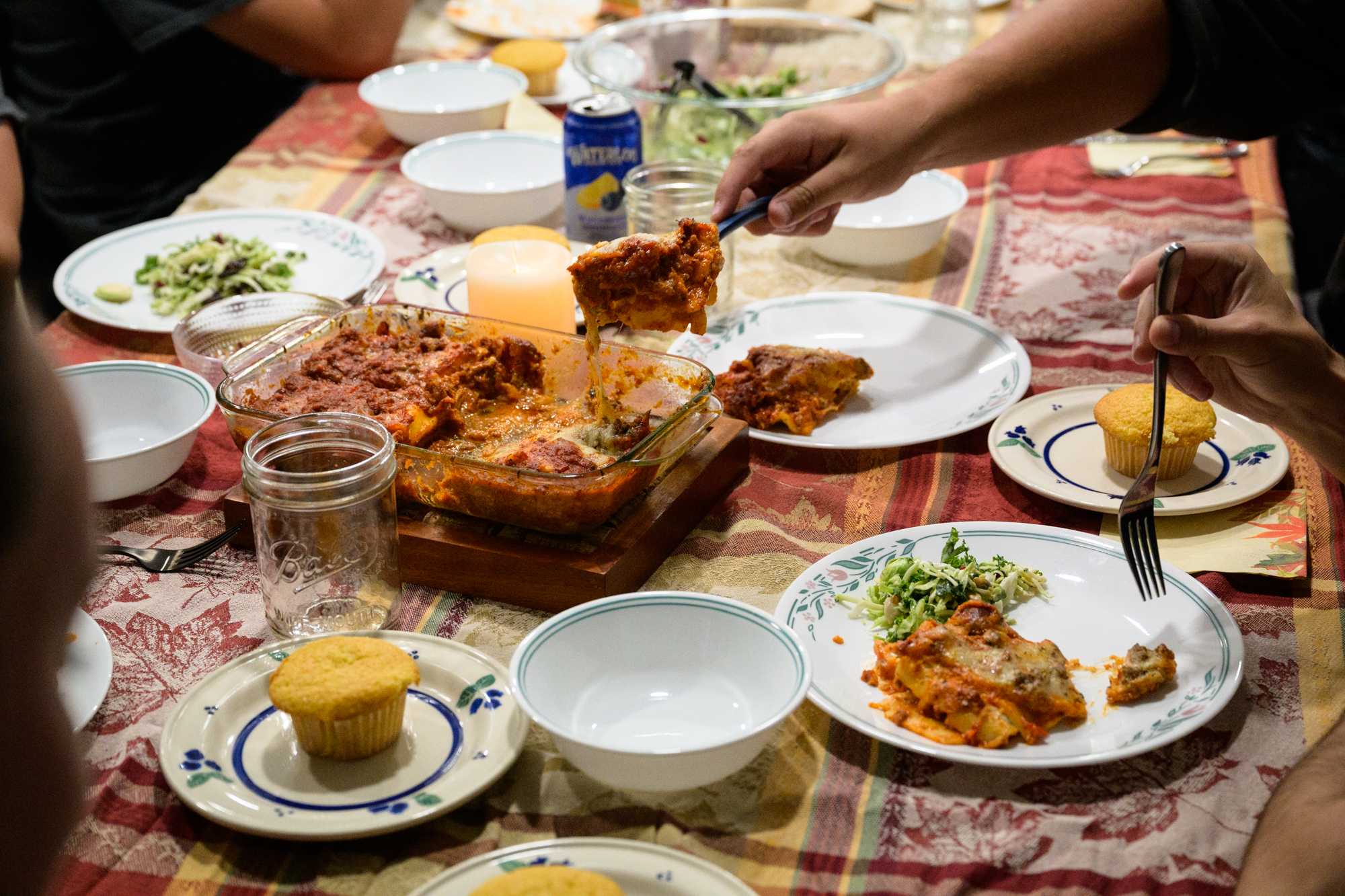 A large pan of lasagna sits between many plates on the table as a hand dishes out a large portion.