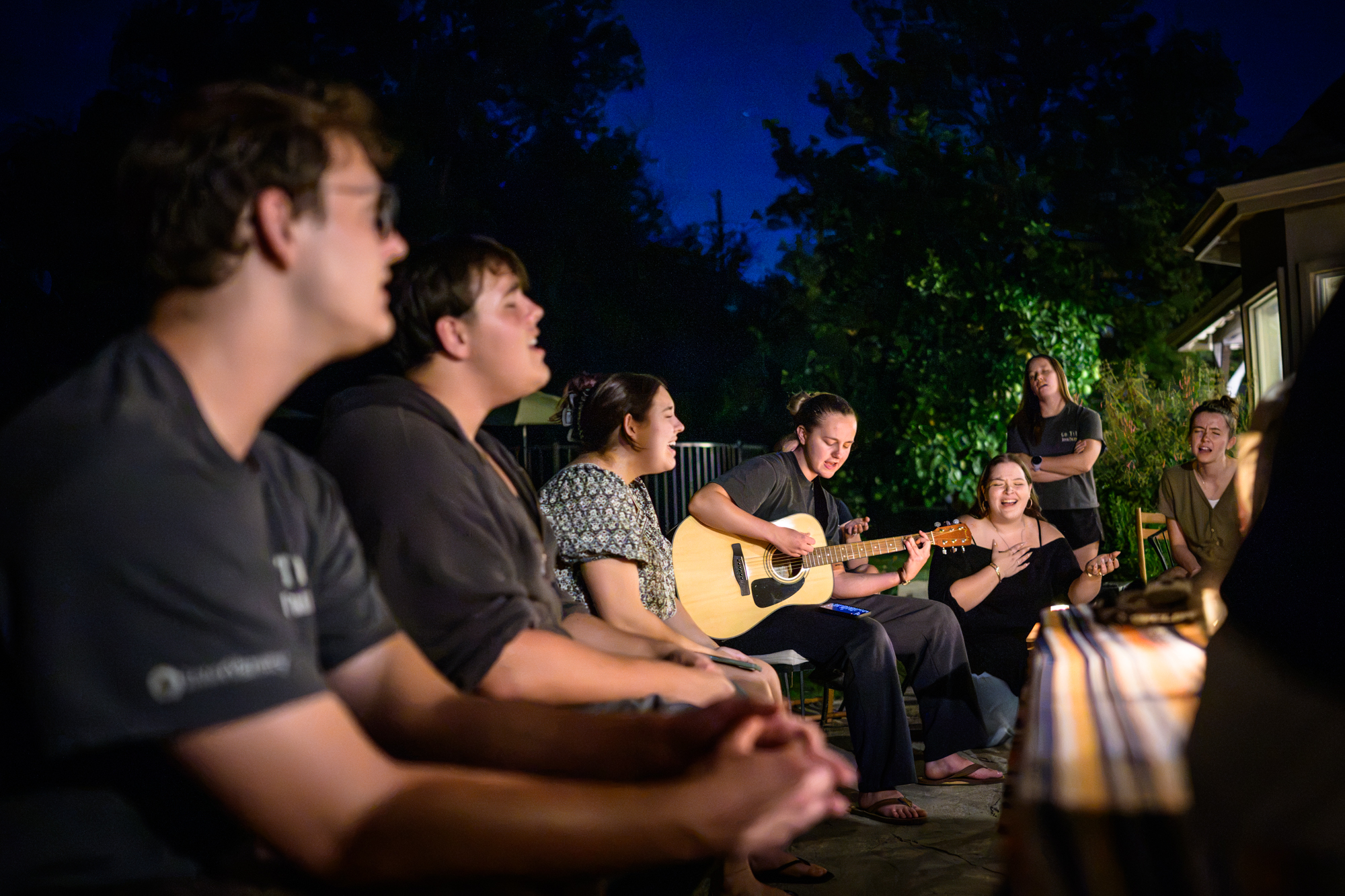 Outside on the back patio under a clear night sky, Clair leads worship with a borrowed guitar as several other students worship with their eyes closed.