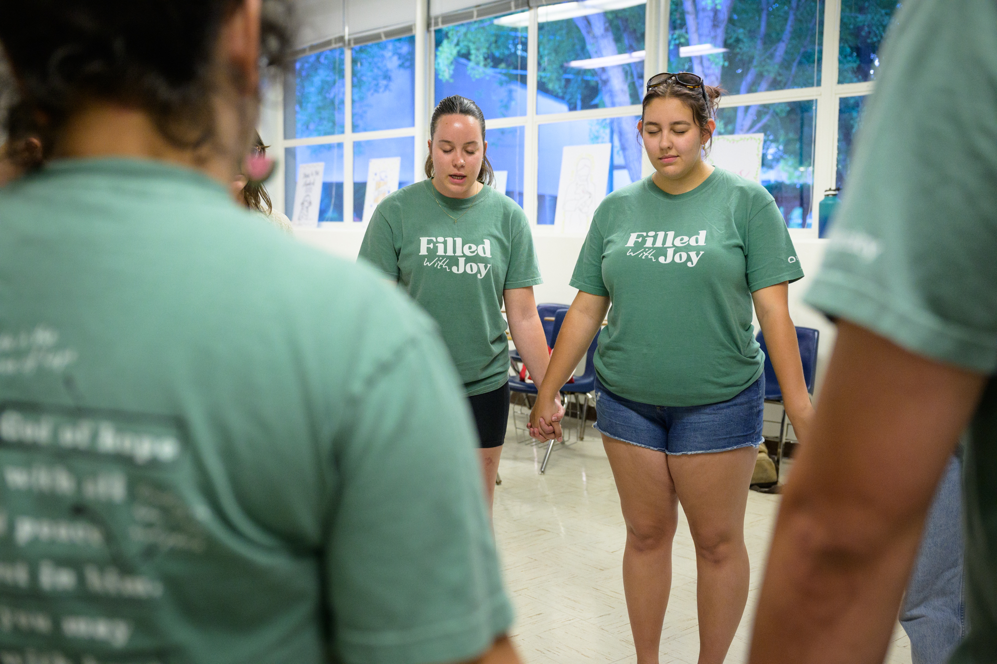 Students that we recognize from all the other photos stand hand in hand praying in a classroom before large group, wearing their chapter t-shirts that read, “Filled with Joy”.