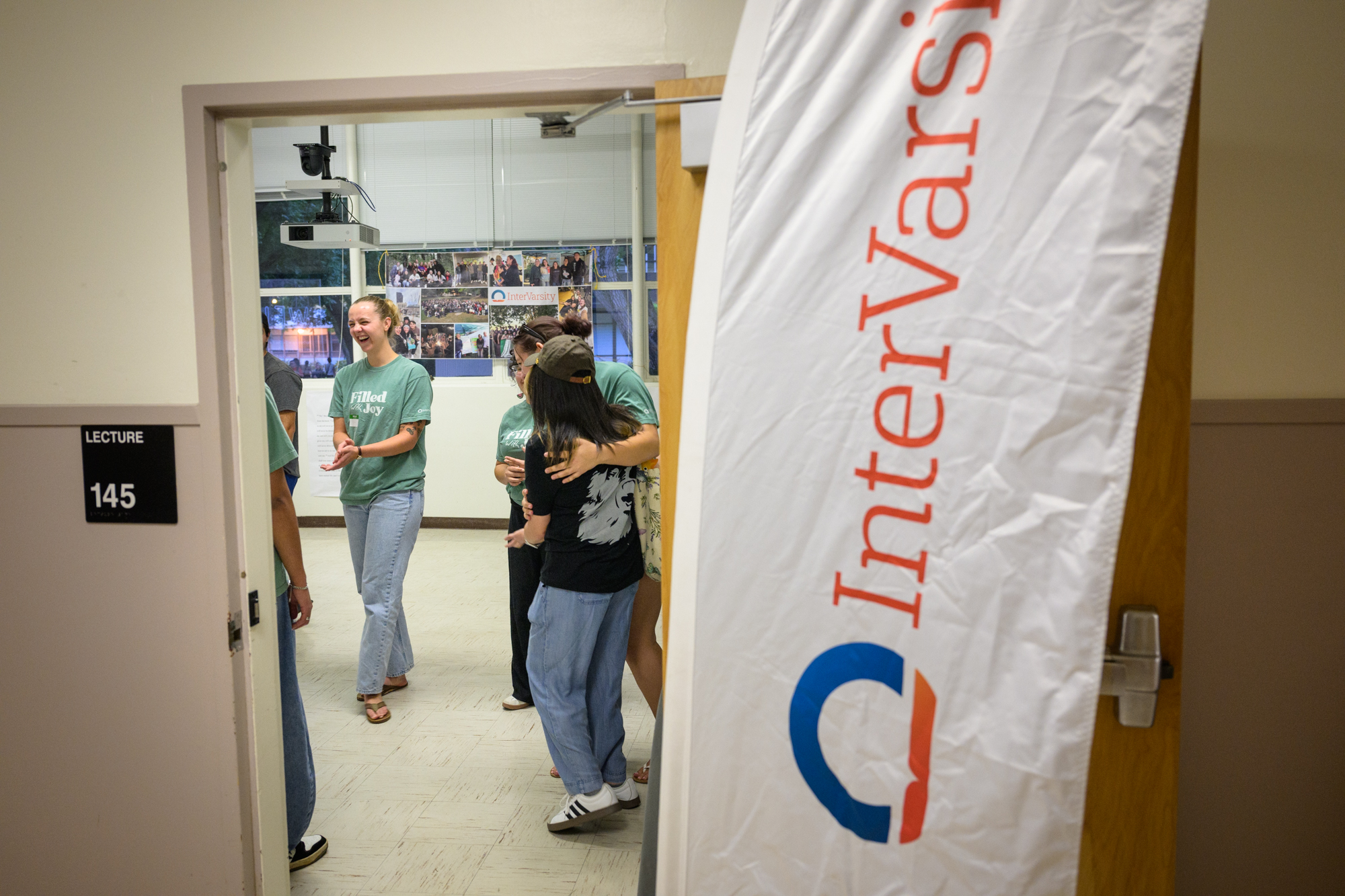 An InterVarsity banner flies outside of a classroom; through the door several students laugh and hug as they see each other.