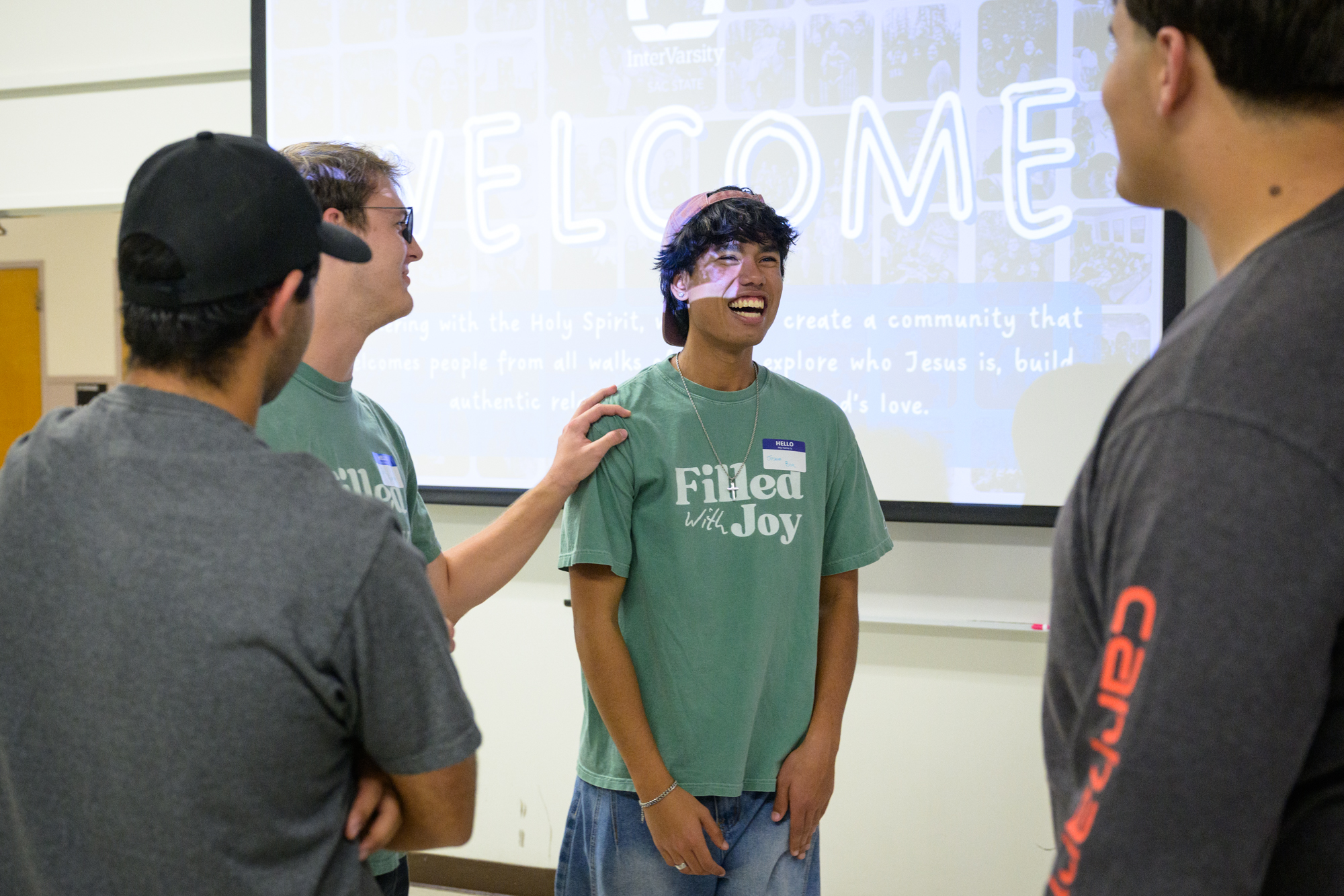 Josh, standing between several other students, laughs as a playful hand grabs his shoulder; a welcome sign appears on the projector screen behind him.