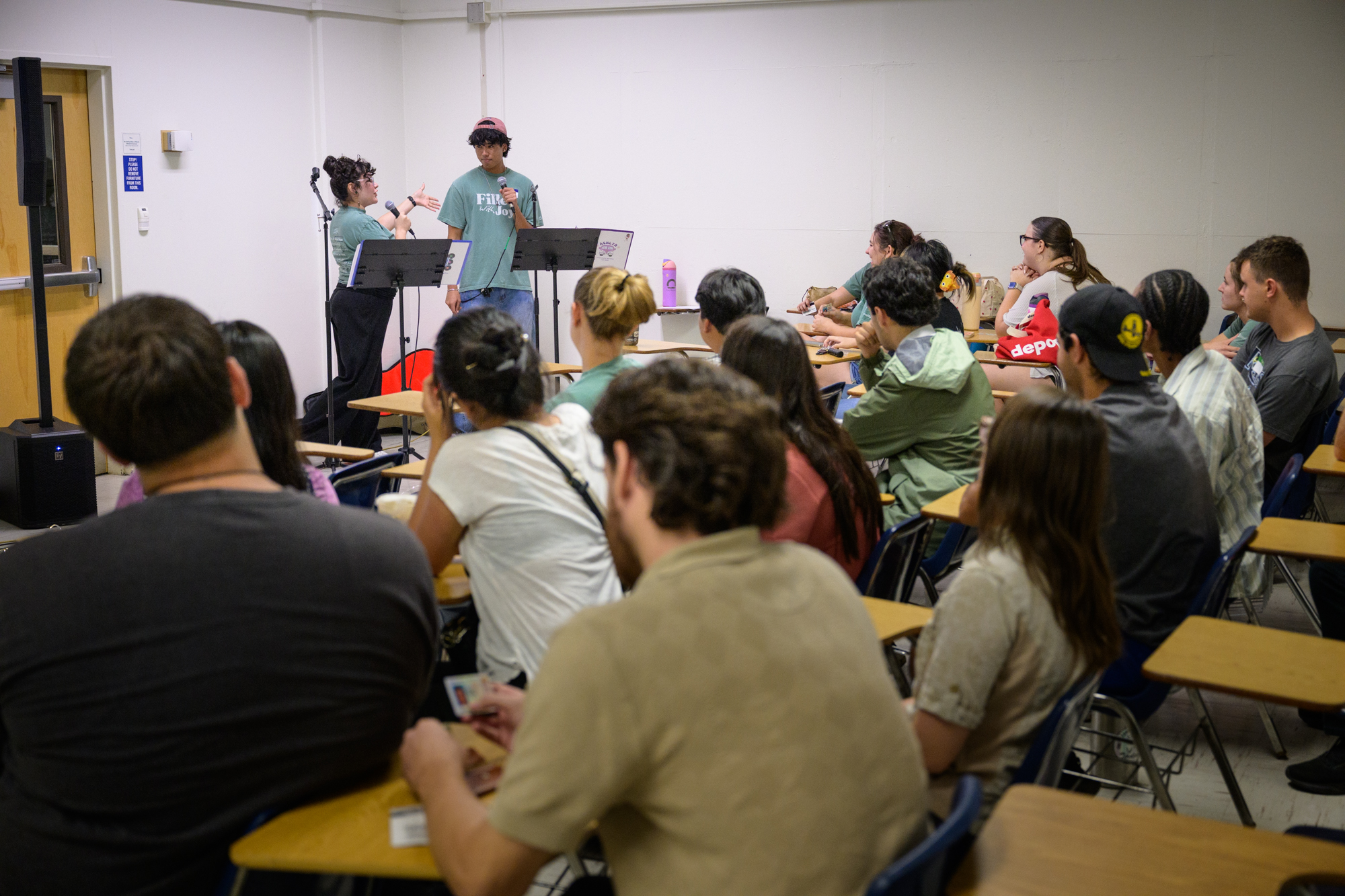 Josh stands up front with a fellow student as they emcee large group; many students sit in desks in front of them.
