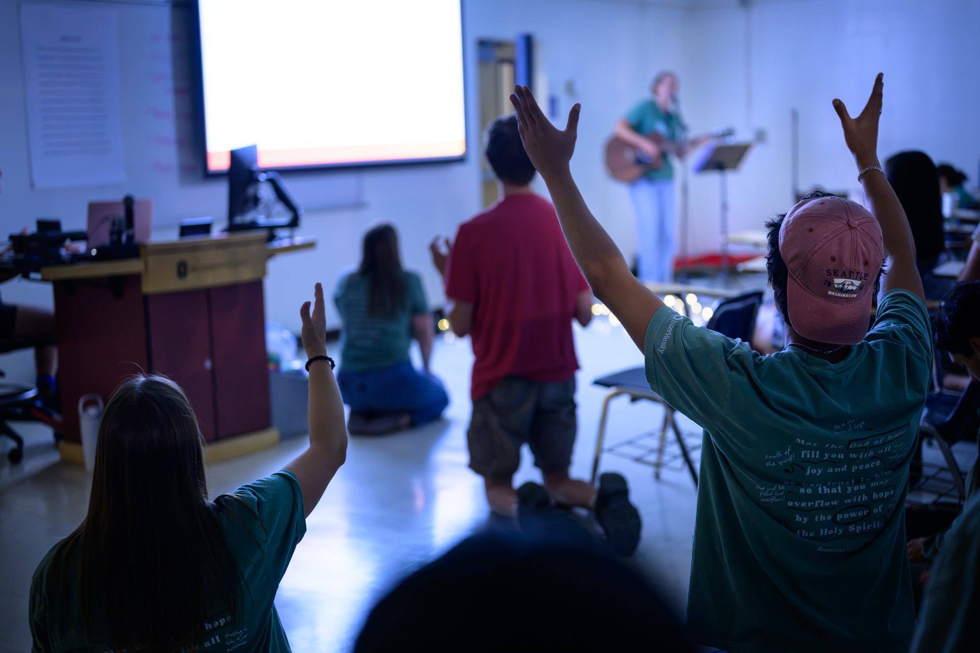 Students are seen kneeling in worship as Clair leads with her guitar in the front of a dimply lit classroom.