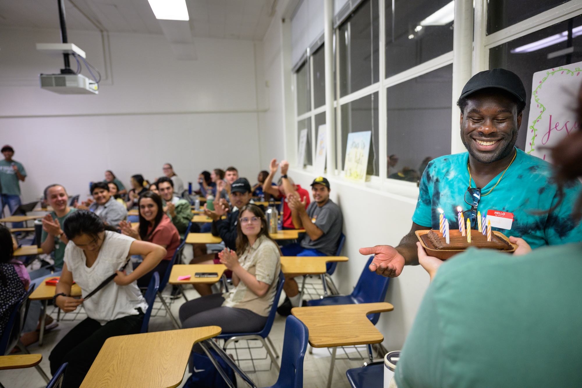 A student smiles as he is presented with a small cake with burning candles; the rest of the large group is seen behind him cheering.