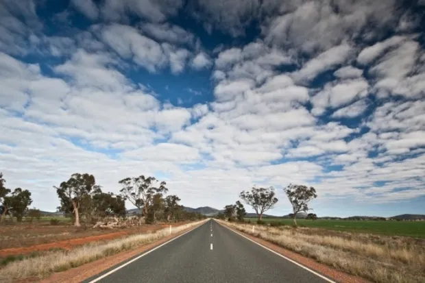 Highway running through rugged landscape with scattered trees and swathes of white clouds