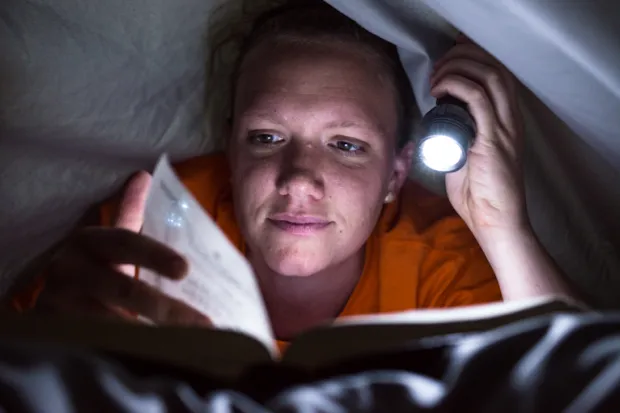 Young woman reading book under covers with flashlight