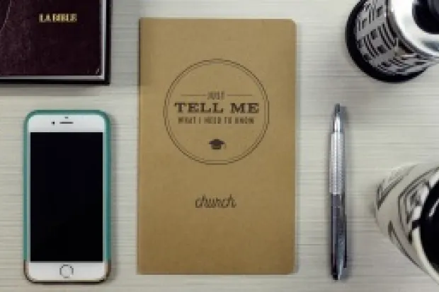 Office desk with phone, pen, Bible, coffee tumbler, and notebook with the words "Just tell me what I need to know about church."