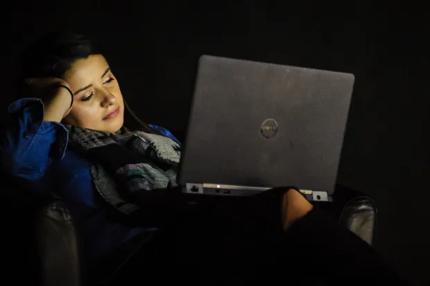 Woman in dark room with laptop glowing on face, looking bored