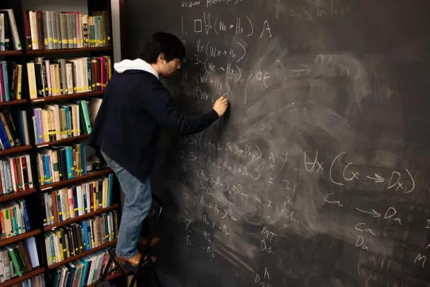 Student standing on ladder writing equation on enormous chalkboard