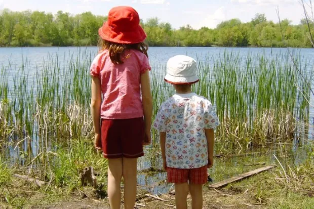 two children, one tall and one short, standing side-by-side and looking out over a lake