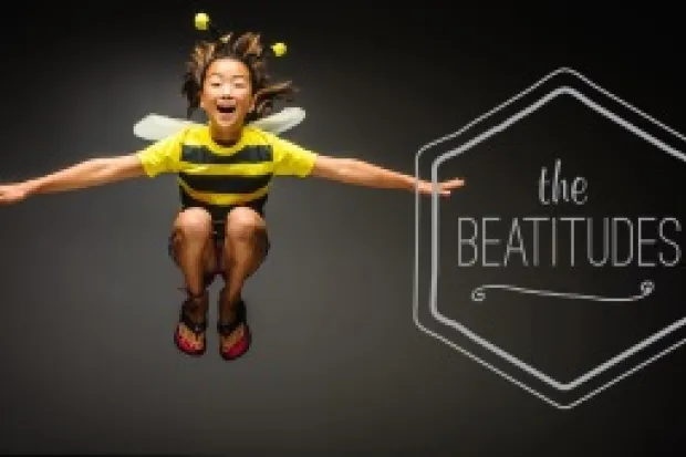 Young child in bee costume in midair, grinning, with the words "The Beatitudes" in the background.