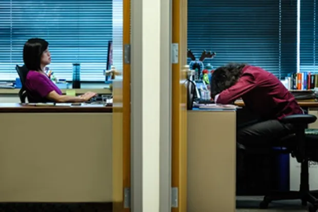 Two offices side by side with doors open. One woman is typing at her computer while the woman in the next office has her head down on the desk.