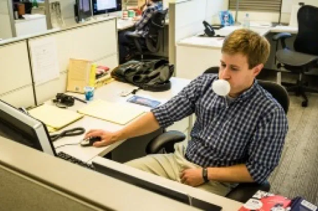 a man sitting in a cubicle blowing a bubble with gum