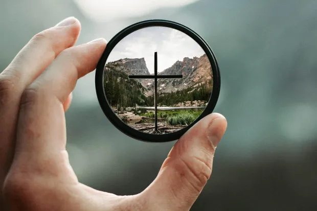 Hand holding camera lens focused on cross with blurry background