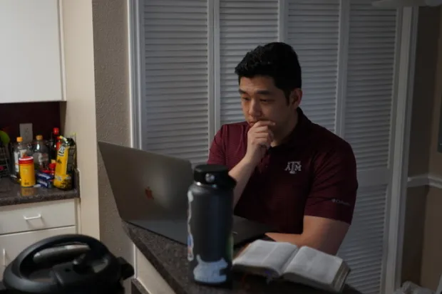 Student reflecting on Scripture, standing at kitchen counter with laptop