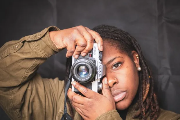 A young black woman holding a camera over her right eye