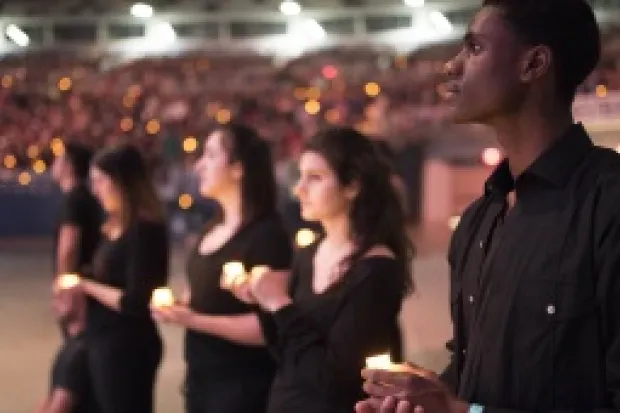 Group of students wearing black holding candles with much larger group in the background in stadium seating