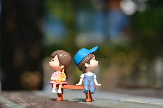 A boy and girl plastic figurine sitting on a tiny park bench and facing opposite ways