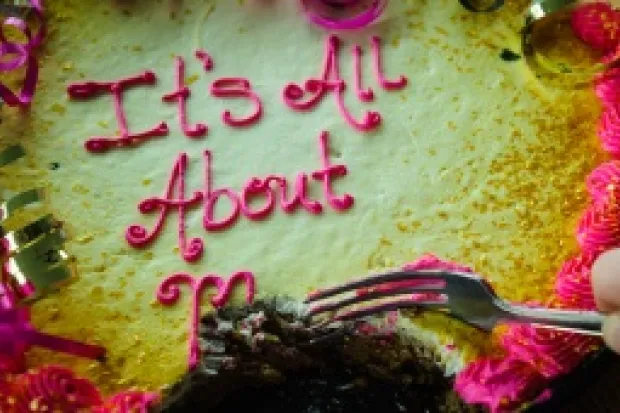 Closeup of pink and yellow cake with "It's All About Me" written in icing. Part of the "Me" has been eaten with a hand and fork poised for more.