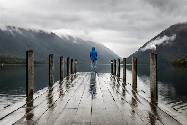 A person standing a long at the end of a rainy dock, facing a lake and distant mountains