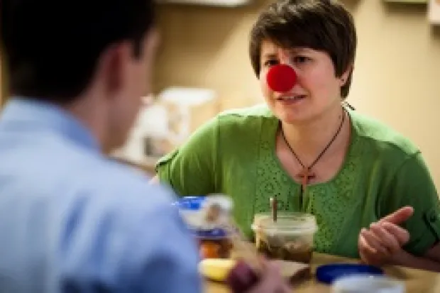 Middle-aged woman wearing red clown nose talking to man across table