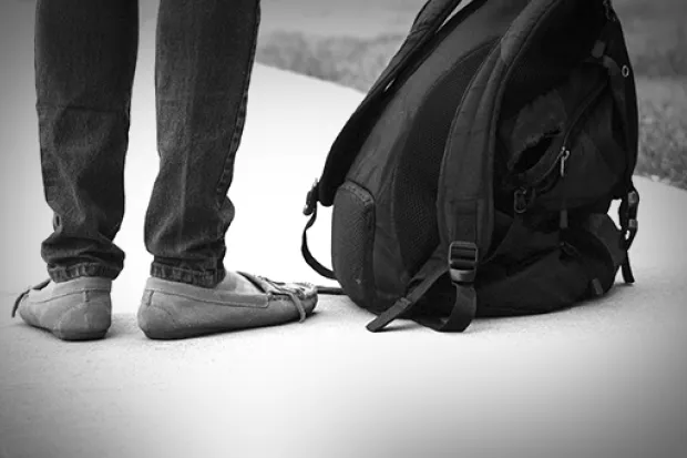 Black and white closeup of person's legs standing on sidewalk next to backpack