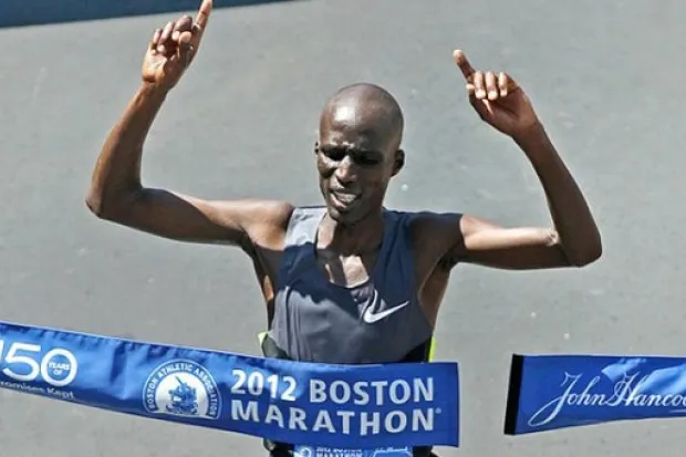 Wesley Korir breaking through tape at Boston Marathon smiling, holding up index finger