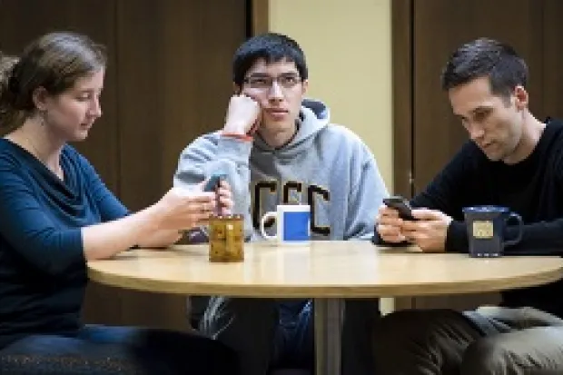 Three students sitting at a table. Two are focused on texting while another is looking bored and left out.