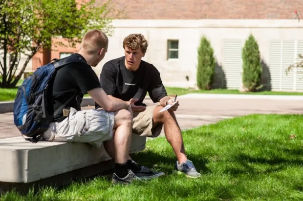 Two male students sitting on a bench on campus talking