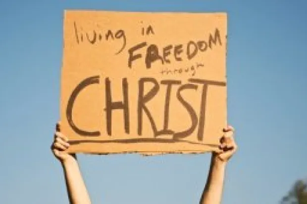 Hands of person holding cardboard sign with the words "Living is Freedom through Christ"