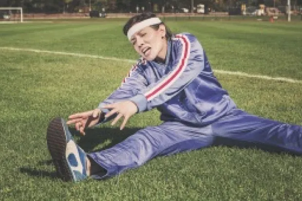 Person on soccer field in bright purple tracksuit grimacing as she stretches