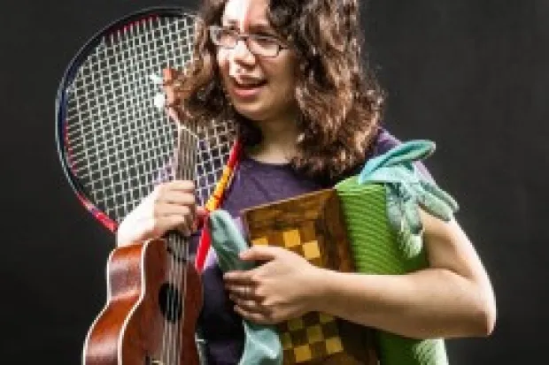 Female college student holding guitar, tennis racket, chessboard, books, and blanket looking overwhelmed