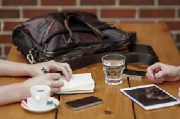 two pairs of hands resting on a wooden table with cups and an iPad on it.