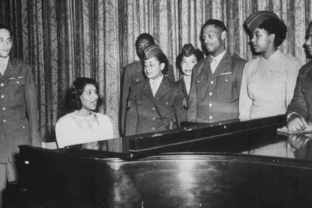 a photo from the 1940's of Black people standing around a piano and singing