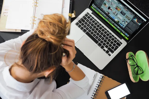 Woman looking stressed, holding her head surrounded by computer and notebooks