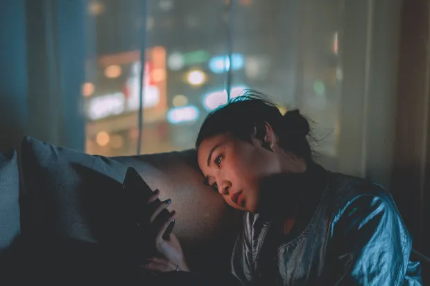 Woman sitting in dark room staring at phone. Window in the background of night cityscape.